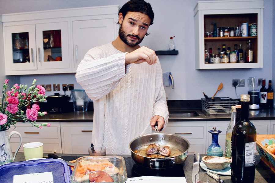 A young man with dark hair and a beard sprinkling salt over a pan with a piece of meat. Next to the pan is a box labeled "kidneys" and a note saying "Enjoy me"