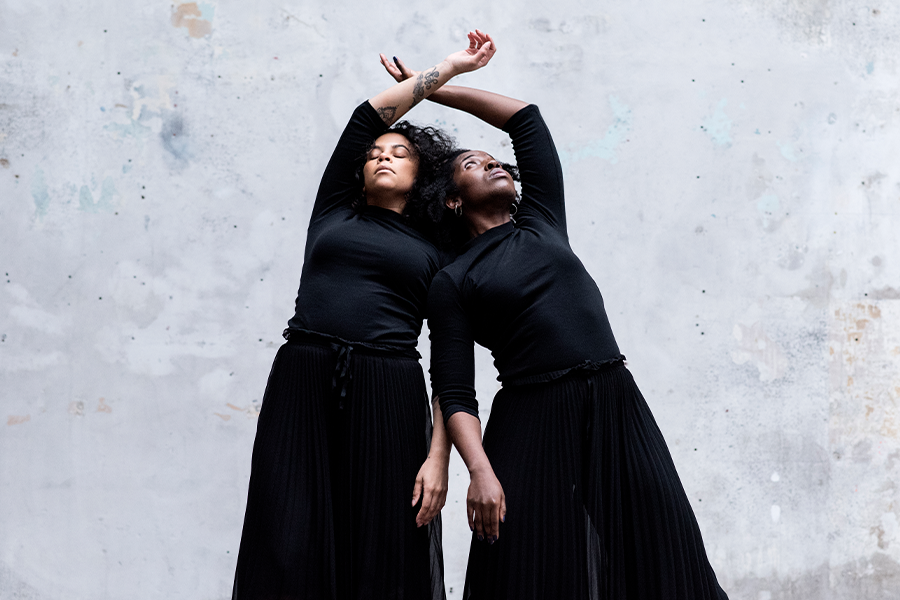 Two people of colour with their eyes closed in a dance pose side by side with their arms crossing above their heads