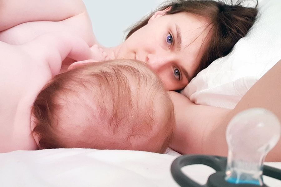 An image of motherhood. A young blue-eyed woman lies on a white pillow in bed, with her baby beside her.