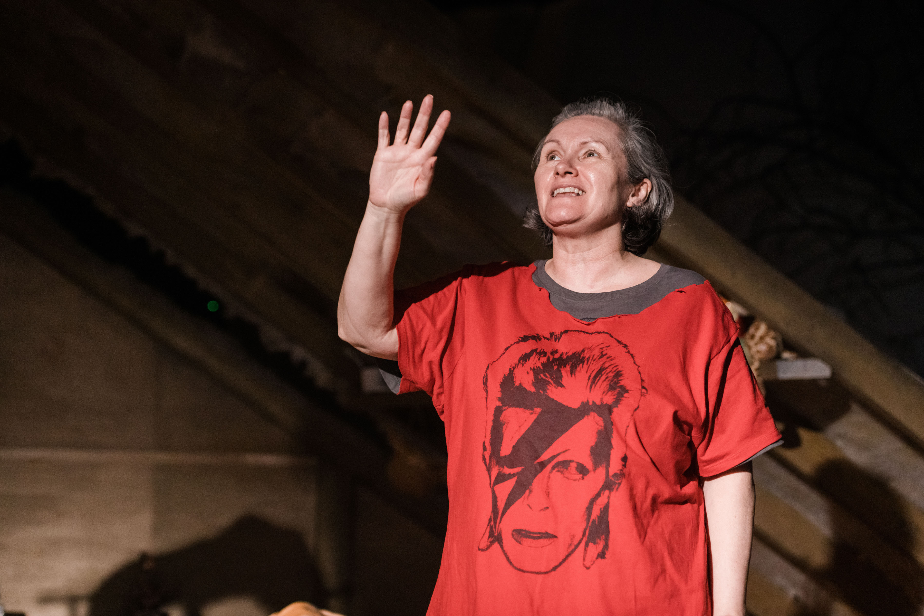 A middle aged white woman dressed in a red Bowie t-shirt, with one arm up
