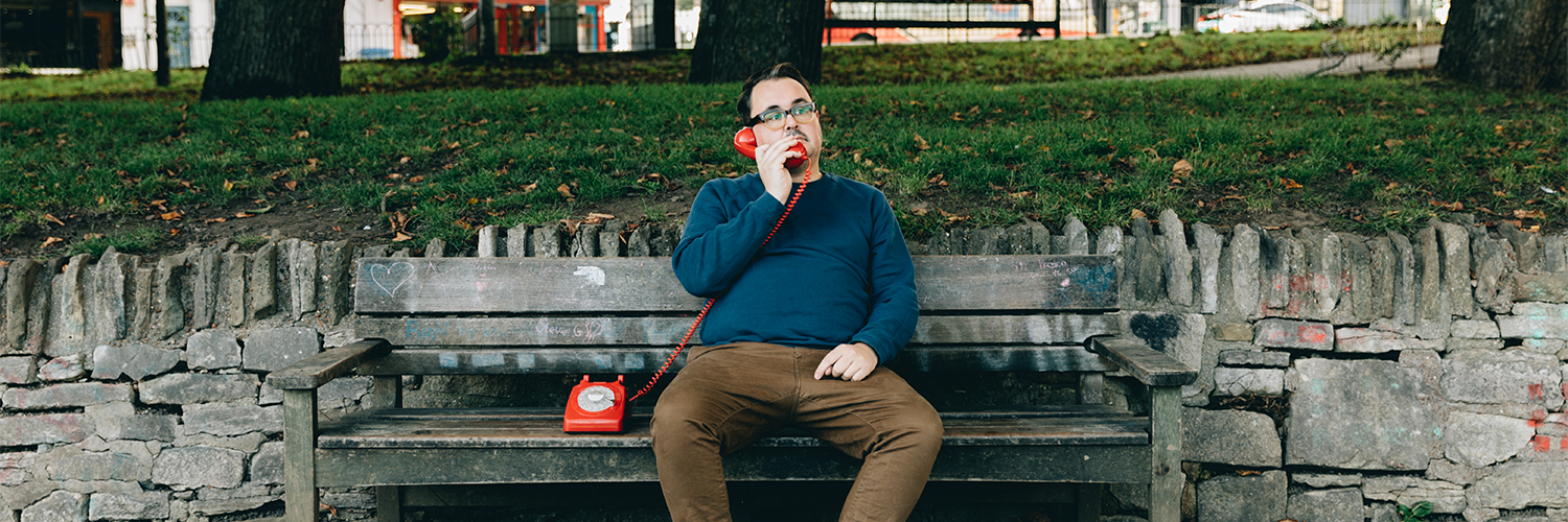 A caucasian person sitting on a bench in the park holding the receiver of a red rotary phone which is on the bench next to them