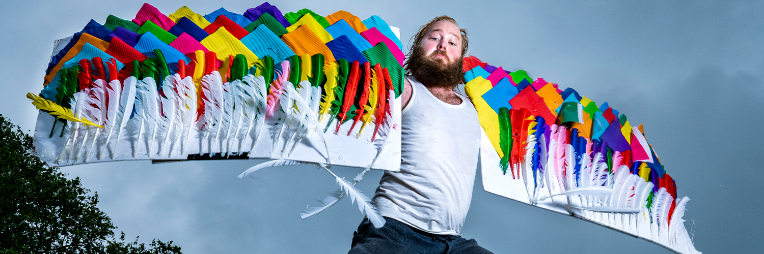 A picture of a caucasian man with a large beard and colourful patchwork wings made of fabric and feathers. He is mid-flight, jumping over a large log with some trees seen in the background