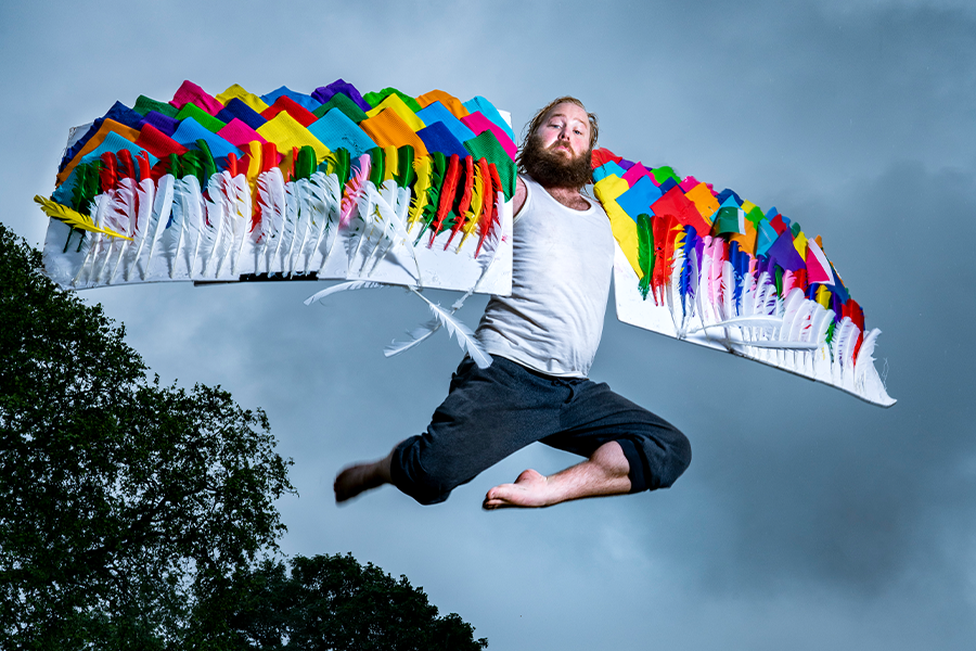 A picture of a caucasian man with a large beard and colourful patchwork wings made of fabric and feathers. He is mid-flight, jumping over a large log with some trees seen in the background