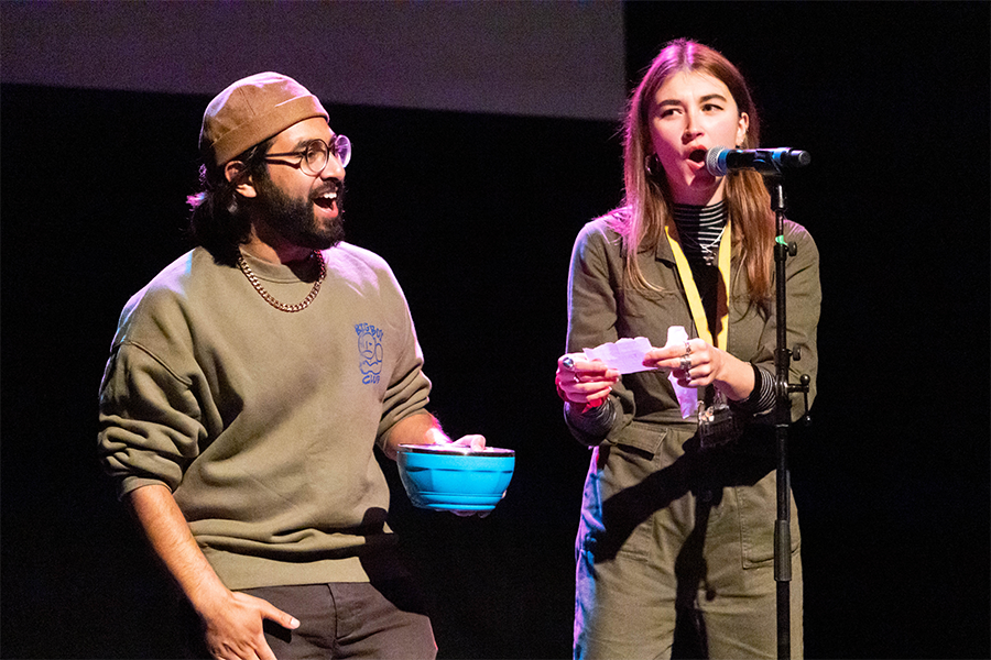 Two performers on a stage in front of a mic. One of them is holding a blue bowl and the other is reading out from a piece of paper that she has taken out of the bowl