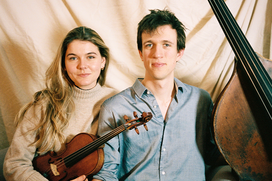 A photograph of two artists looking directly into the camera. One of them is holding a violin and next to the other is a base.