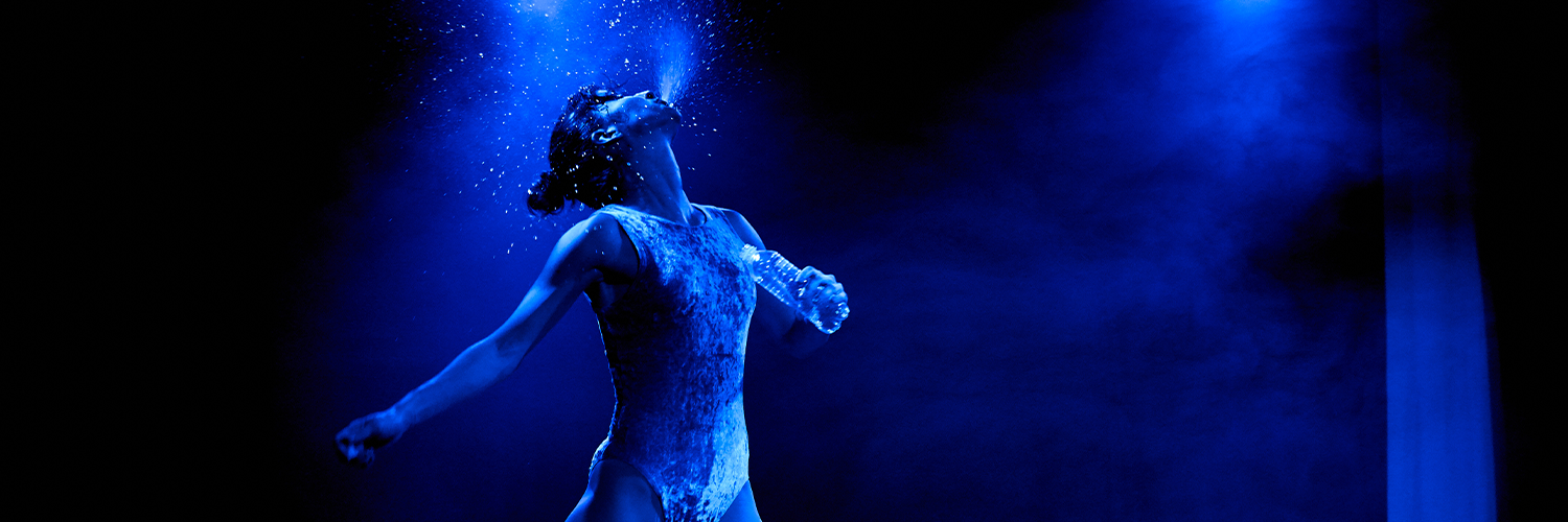 A performer dressed in a body suit with their head back spitting out water and holding a bottle of water in one hand