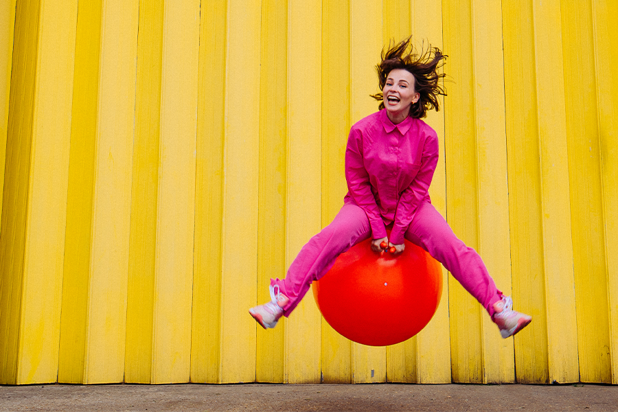 A young caucasian woman in bright pink jumpsuit bouncing on a red junmping ball midair in front of bright yellow background