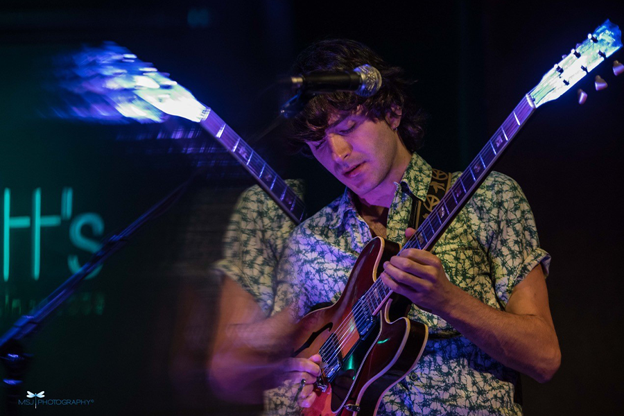 An image with a slight mirrored effect of a man in a short sleeved shirt playing an electric guitar on stage
