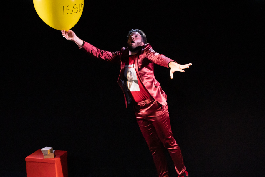 A caucasian man in a shiny red suit and bright red shoes caught midair on stage while holding a yellow balloon that has "Daddy Issues" written on it