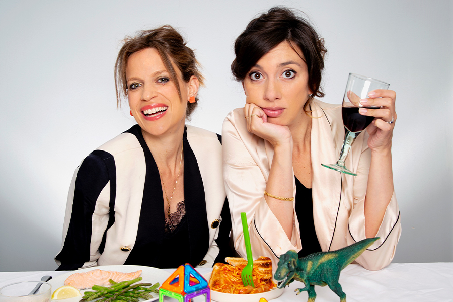 A photo of two women sitting side by side at a table that has kid's toys and food on it. One of the women is drinking a large glass of red wine. Both are looking at the viewer.