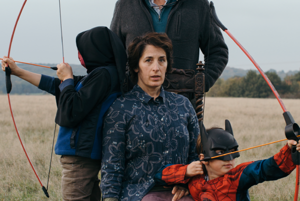 A portrait of a woman in a field. She is surrounded by two small children carrying toy bows and arrows. Behind her stands a man whose face we don't see