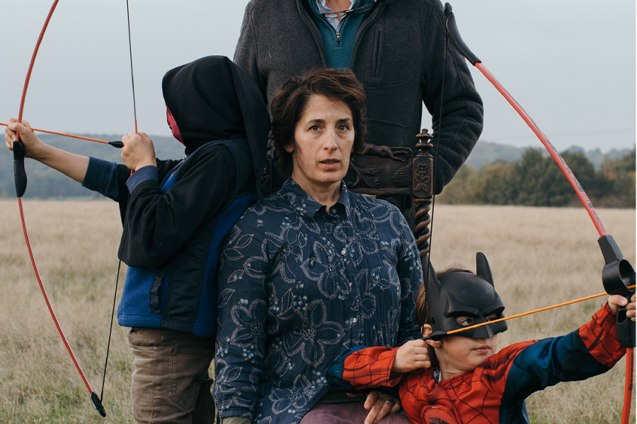 A portrait of a woman in a field. She is surrounded by two small children carrying toy bows and arrows. Behind her stands a man whose face we don't see