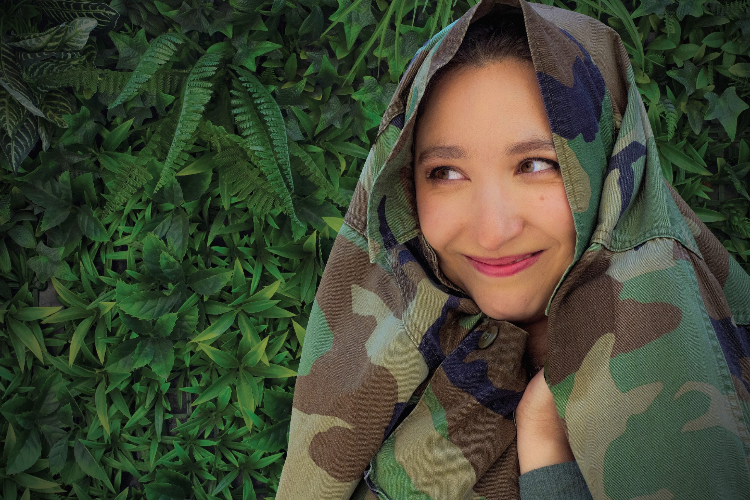 A young woman grins looking to one side in front of a push, and with a camoflage jacket over her head. Themes of the climate crisis.