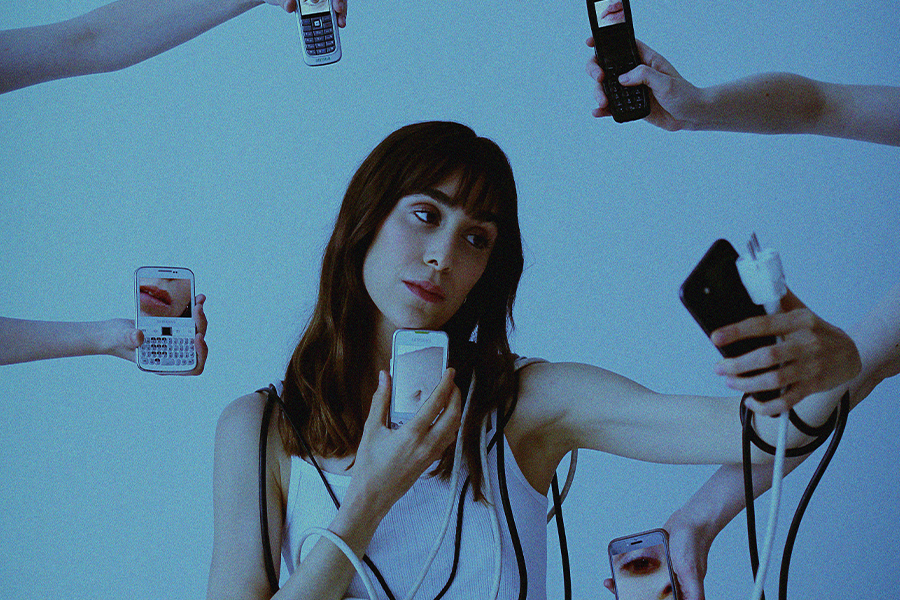 A woman sits taking a selfie whilst wrapped in wires and cables and various floating phones surrounding her.