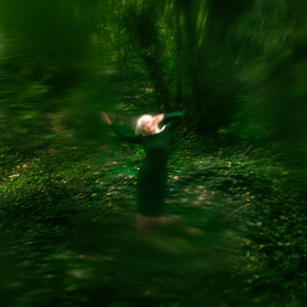 A woman in a green dress stands in the middle of a green landscape, surrounded by trees. Her arms wave in the air above her as she appears to sway. The images is blurred and slightly distorted.