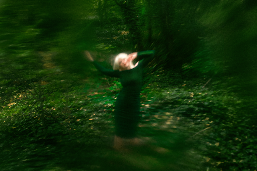 A woman in a green dress stands in the middle of a green landscape, surrounded by trees. Her arms wave in the air above her as she appears to sway. The images is blurred and slightly distorted.