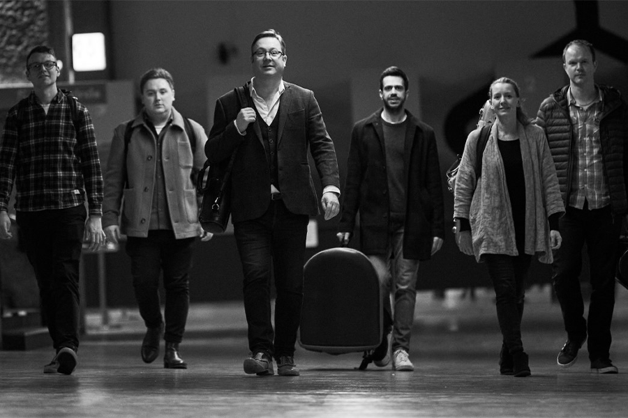 Black and white image of six musicians walking towards the camera with their instruments in hand.