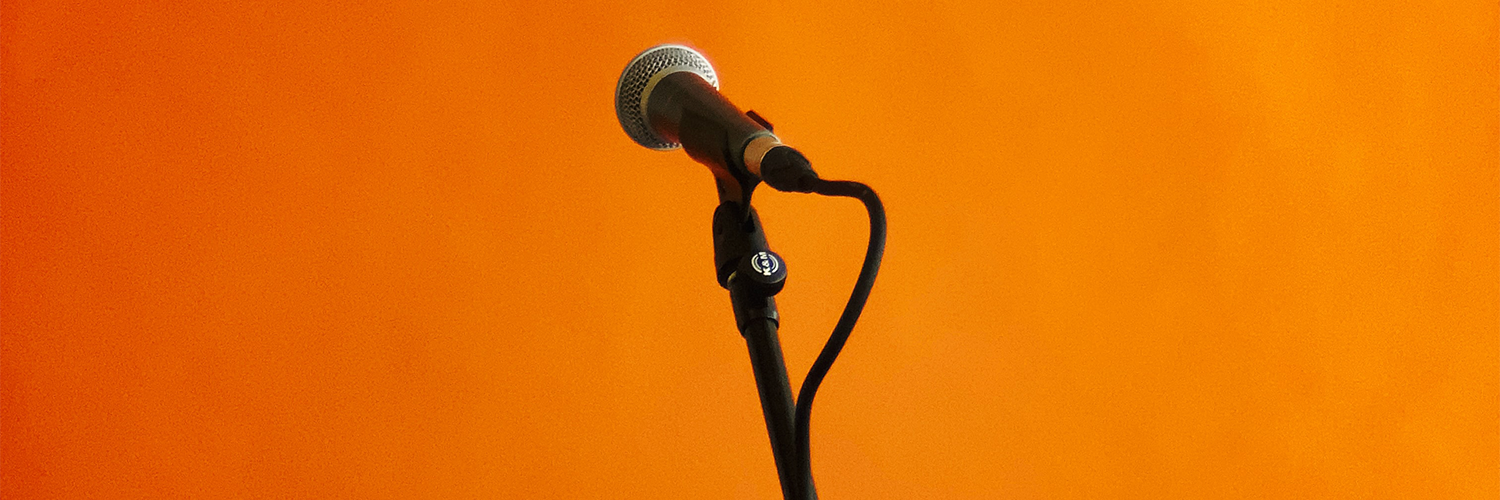 A lone microphone on a stand in front of an orangey red background, with a caberet theme