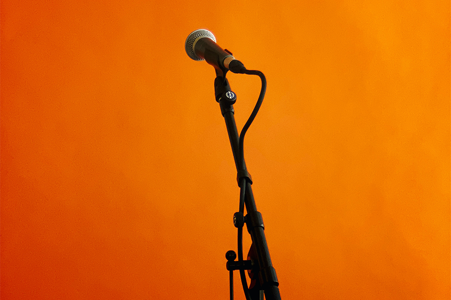 A lone microphone on a stand in front of an orangey red background, with a caberet theme
