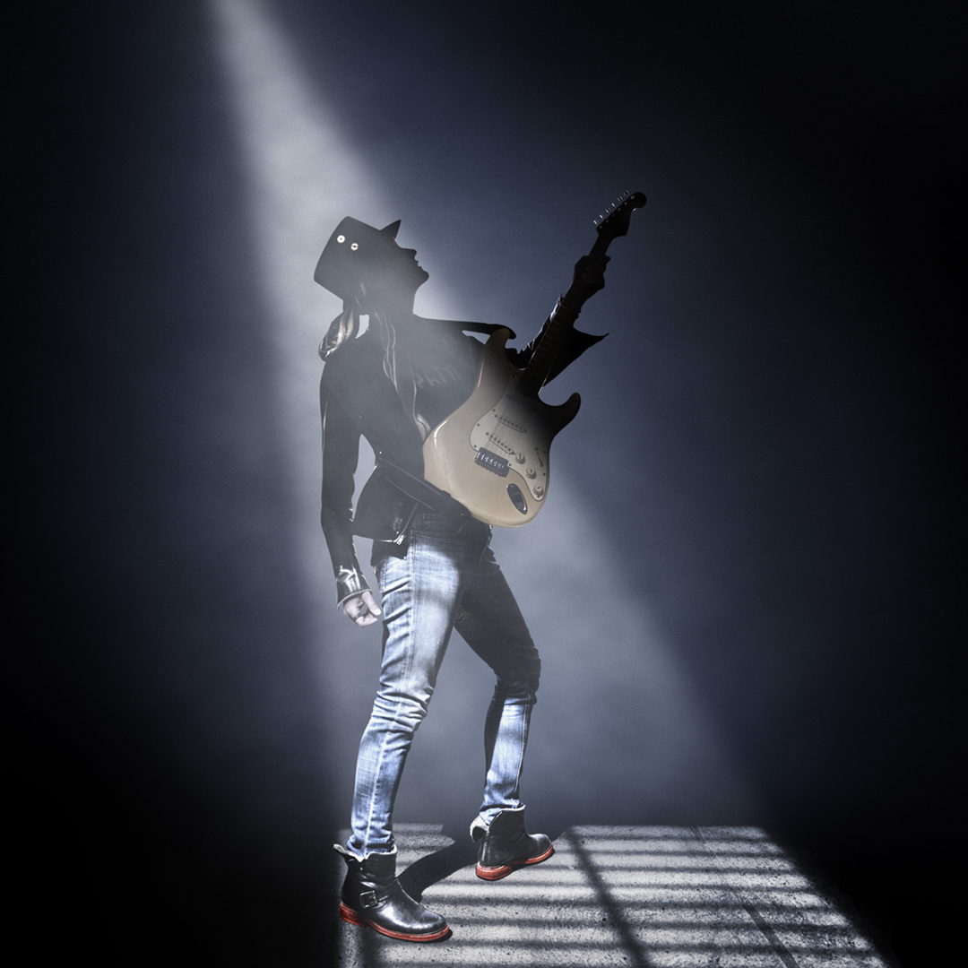 A woman with an electric guitar stands beneath a spotlight on a smoky stage, lead image for Shutters: A Lesbian Rock Opera.
