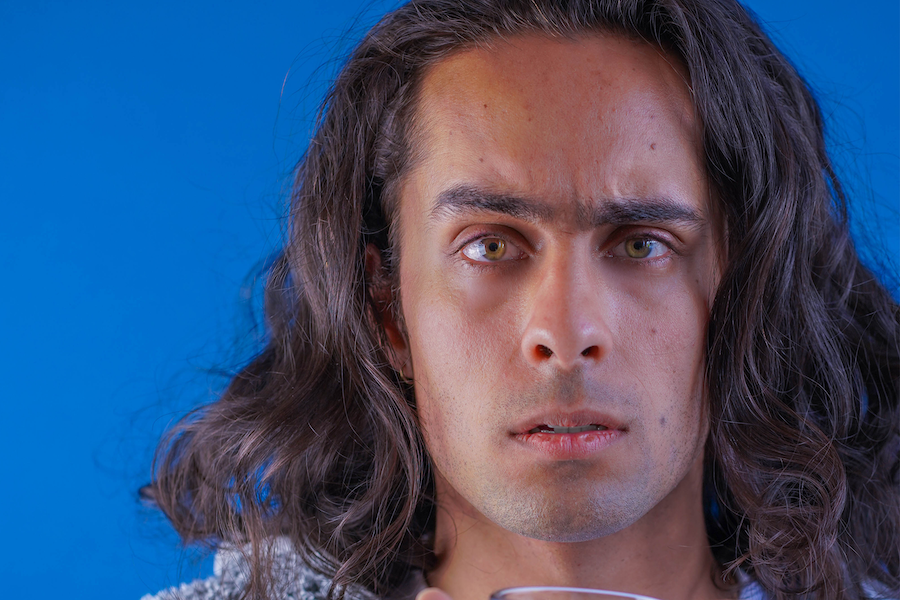 A portrait of a frowning man with long hair holding a cup of tea in a transparent glass, against a blue backdrop.