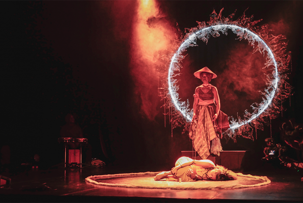 A production shot for Myth Refreshed, featuring a person curled up in fetal position in the centre of the stage and another person hovering above them surrounded by a circular structure with led lights and plants