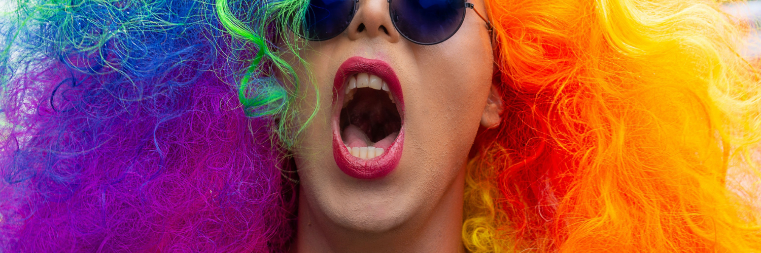 Close up image of a woman in a bright rainbow-coloured wig and sunglasses with her mouth open.