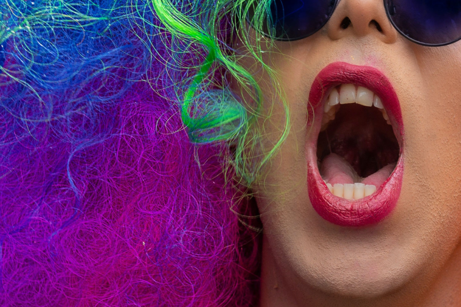 Close up image of a woman in a bright rainbow-coloured wig and sunglasses with her mouth open.