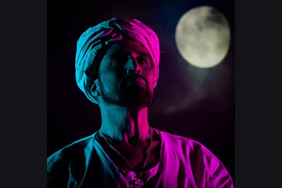 A south asian man wearing a turban is lit in half pink and half blue whilst the projected moon looks over him