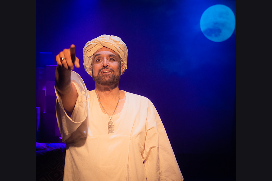 A south asian man wearing a turban and tunic points outwards into the audience. The stage is dimly lit in blue with the moon projected into the right corner