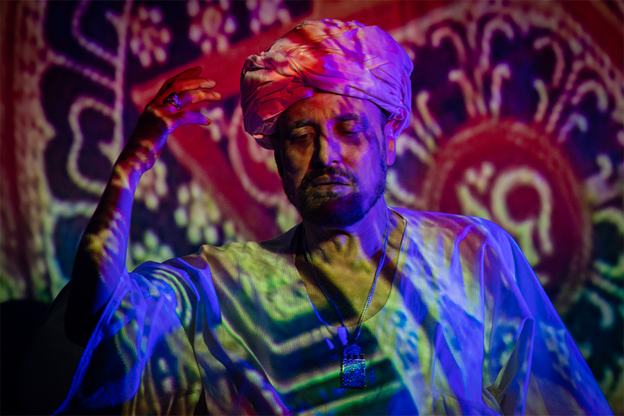 A man wearing a turban holds his hand in the air whilst south asian patterns are projected onto the wall covering his body