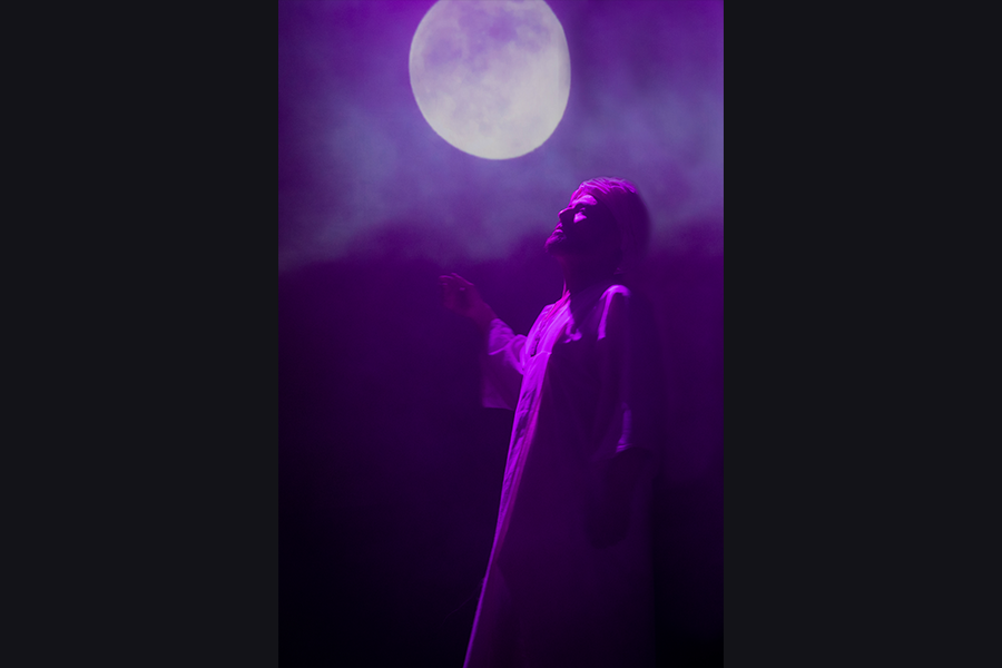 A south asian man wearing a turban and tunic holds his arms open looking to the sky where there is a moon projected onto the stage