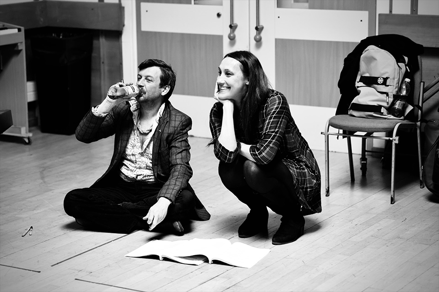 Two people on the floor at the Shutters rehearsal. The woman is crouching looking up smiling and a man is drinking water crossed legged on the floor.
