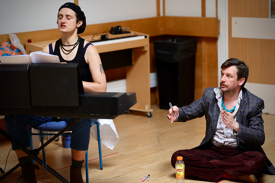 Two people in the shutters rehearsal room. One on the piano and the other on the floor holding a pen mid-explanation