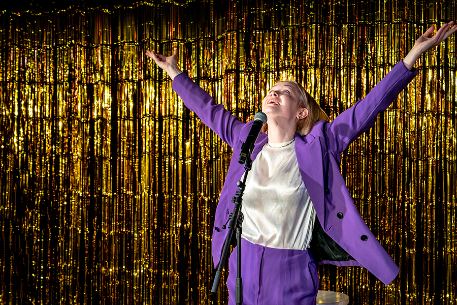 A woman wearing a purple pantsuit holds her arms wide open in the air facing her head towards the ceiling. The backdrop is a shimmery gold.