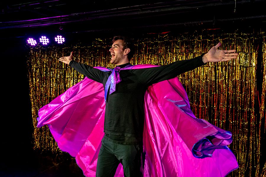 A man wears a pink superhero like cape, holding his arms wide. The backdrop is a shimmery gold.
