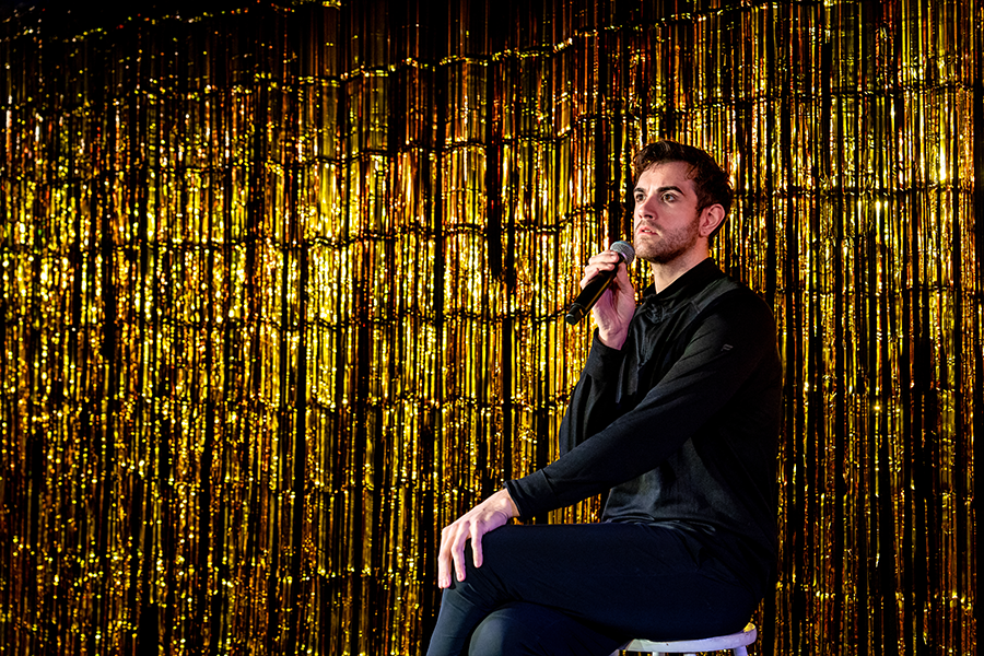A man sits cross legged on the stool with a mic in his hand. The backdrop is a shimmery gold.