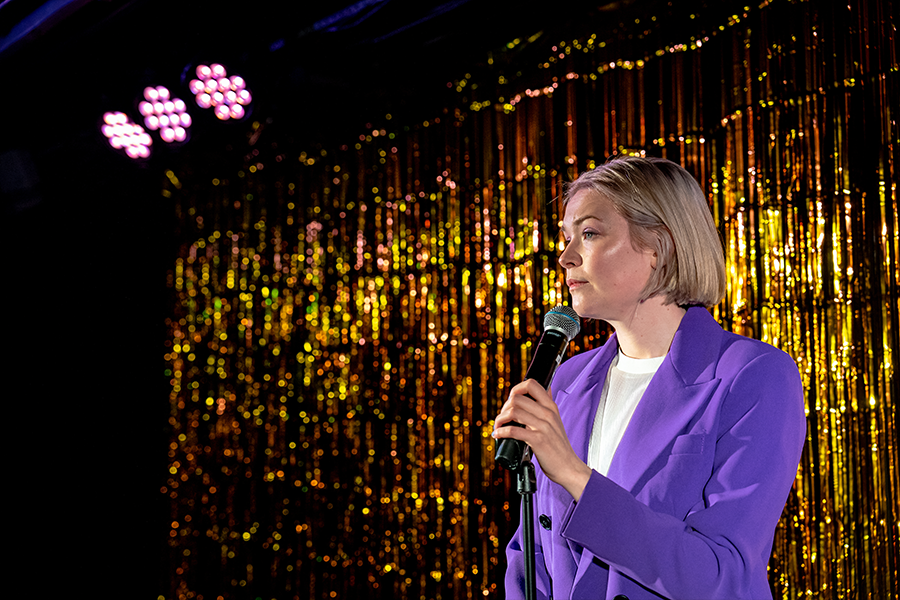 A woman with short blonde hair holds a microphone, wearing a purple blazer. The backdrop is a shimmery gold.
