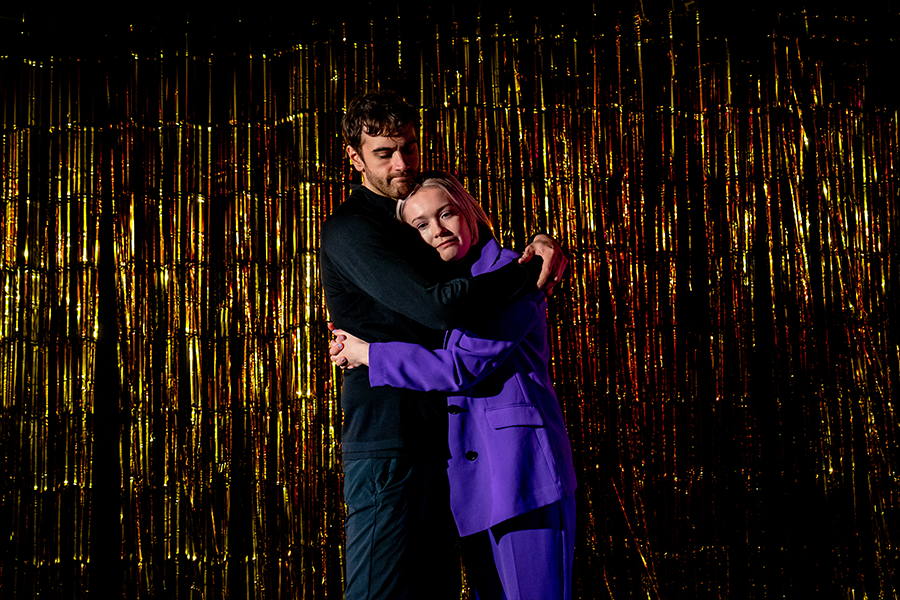 A man and woman hug on stage. The man wears all black and the woman wears a purple pantsuit.
