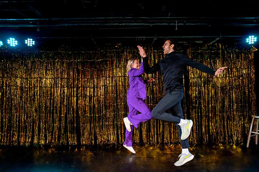 A man and woman are dancing chaotically on stage, skipping mid air and High-fiving.