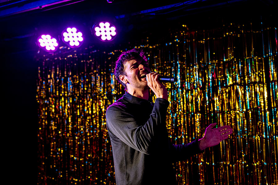 The man is singing into the microphone with strained passion. The backdrop is a shimmery gold.