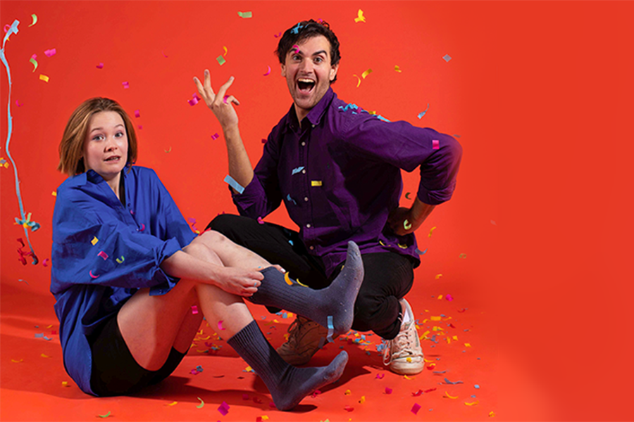 Plain Heroines, the duo. One woman is sat on the floor awkwardly adjusting her socks. Whilst a man crouches next to her confidently hand on hip throwing confetti.