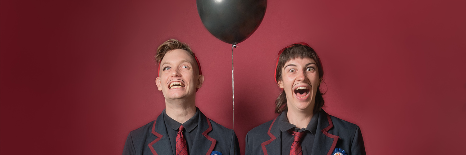 Two performers dressed in drag appearing as young school boys wearing black blazers and red ties. They both look excited with grins and mouths open. A black balloon floats between them.
