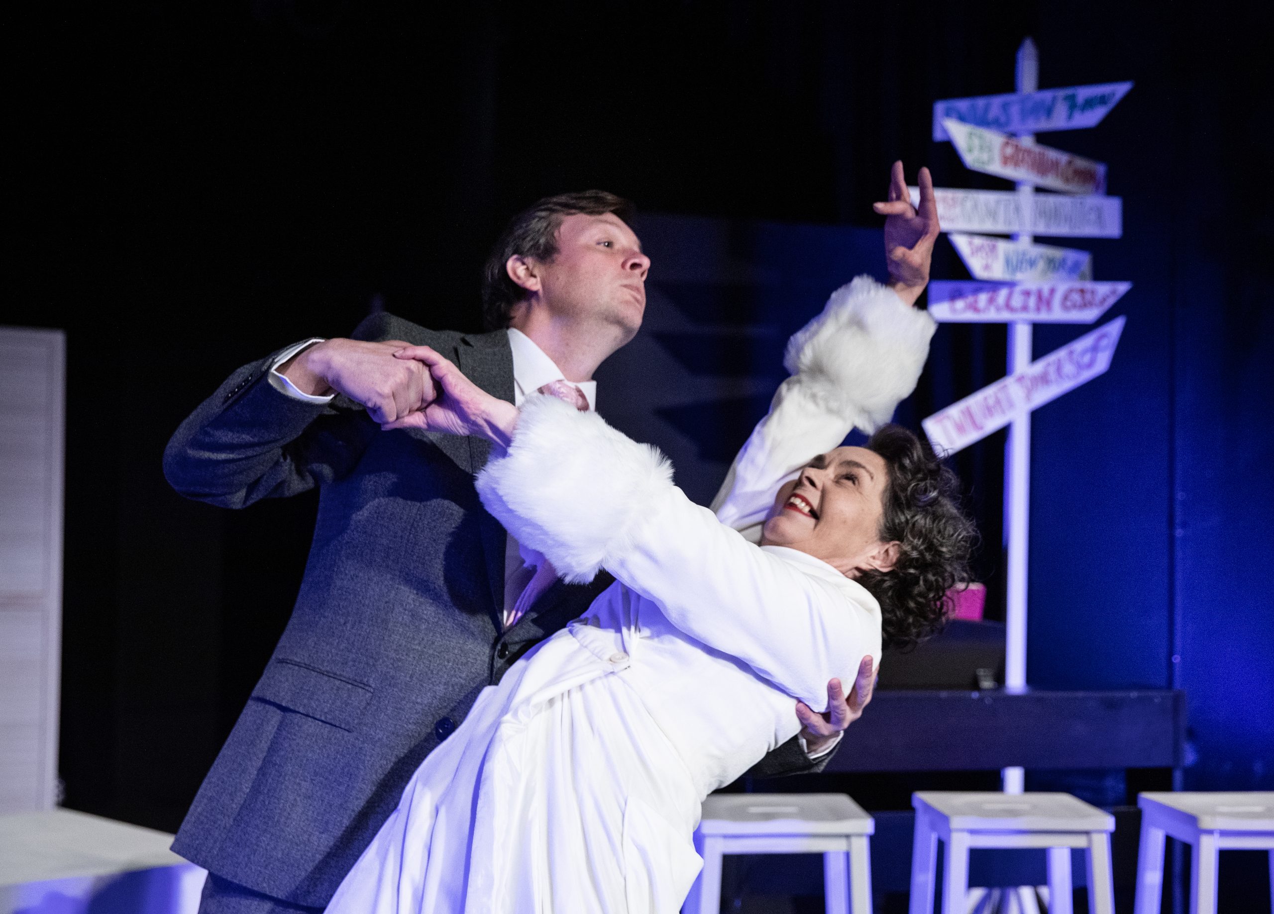 A man in a suit and a woman in a white coat with furry sleeves are dancing, he is dipping her backwards as she holds out her arm