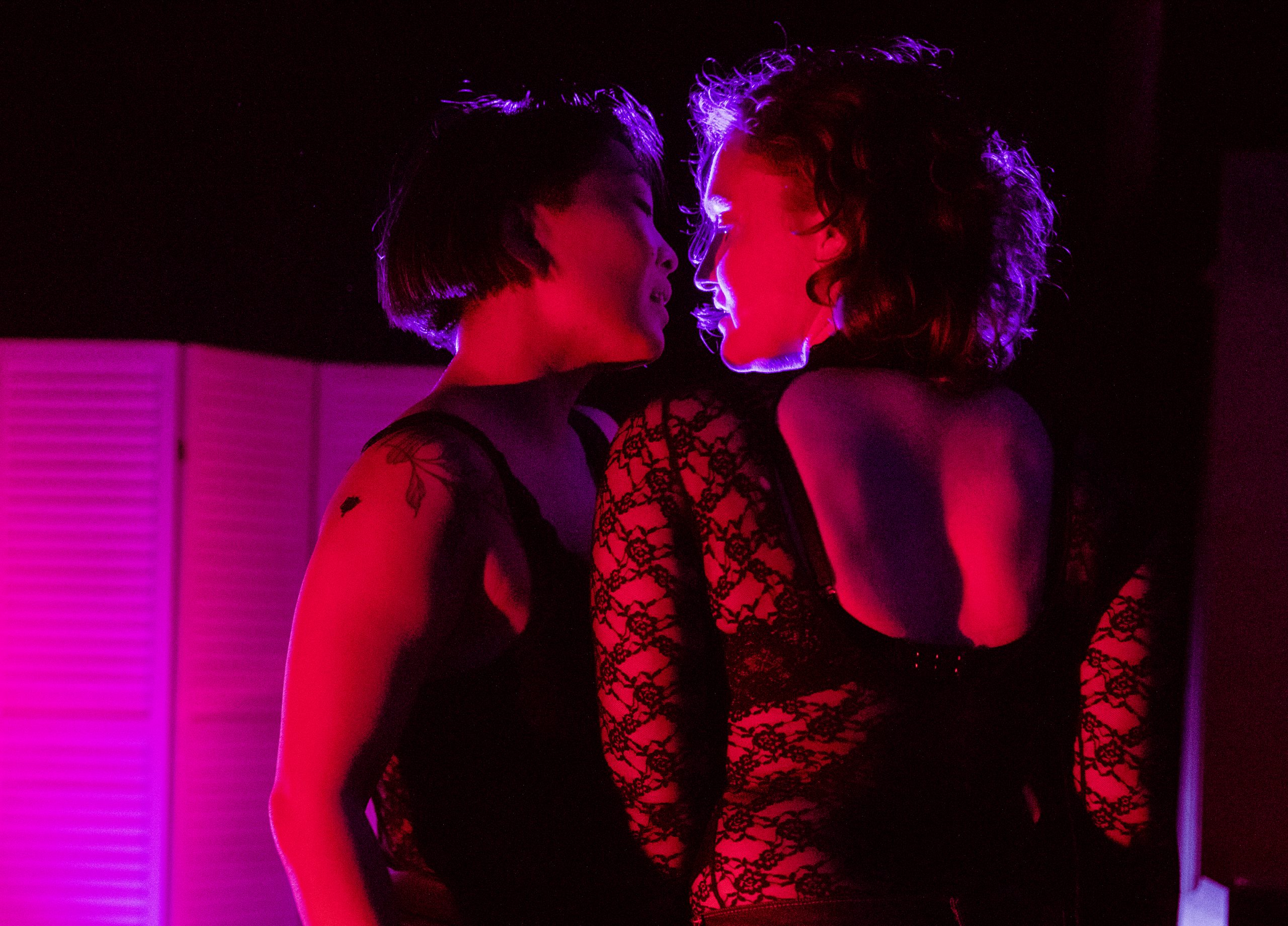 Two women lit in red and pink hold their faces close to one another almost kissing