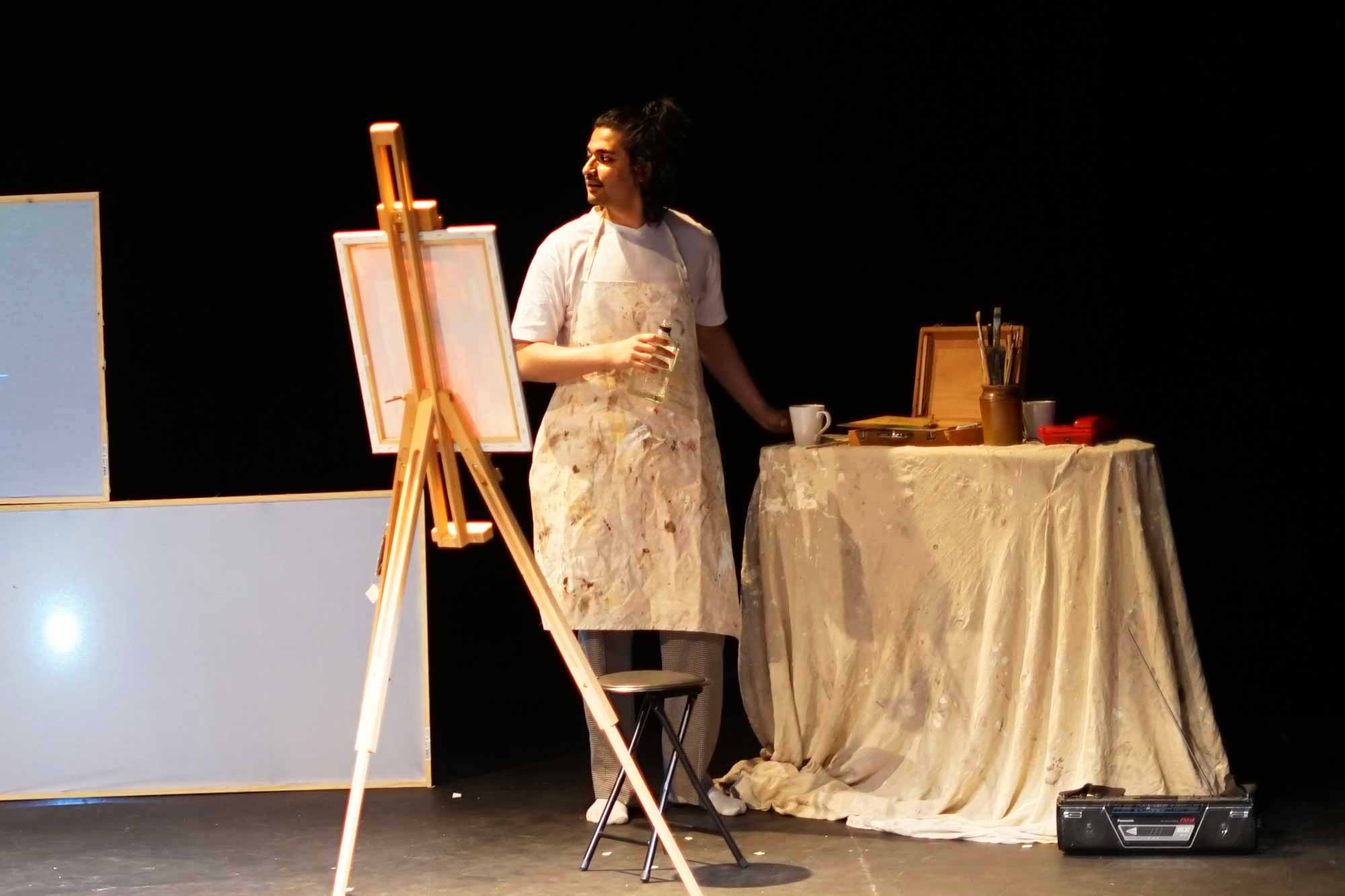 A male artist wearing an apron is looking to the side of his easel by his table of paints