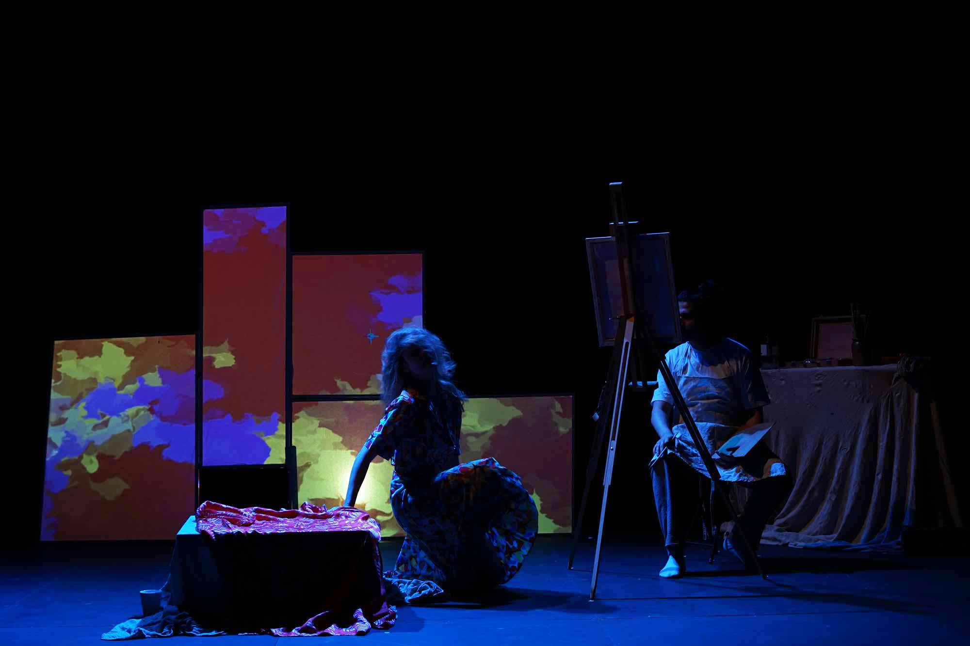 four screens stacked on top of each other creating a geometric effect. With splatters of colours from red, to yellow to purple. A Woman is crouching close to the floor as a male artist paints on his easel.
