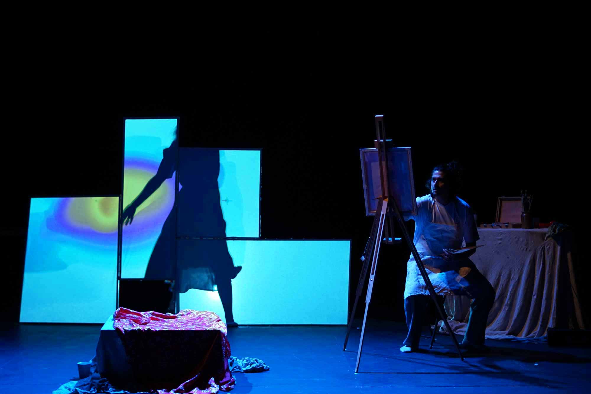 four screens stacked on top of each other creating a geometric effect. The screen shows vibrant colours and auras of turquoise, blue, yellow, orange and purple. A woman is moving in front of it while the male artist is painting her.