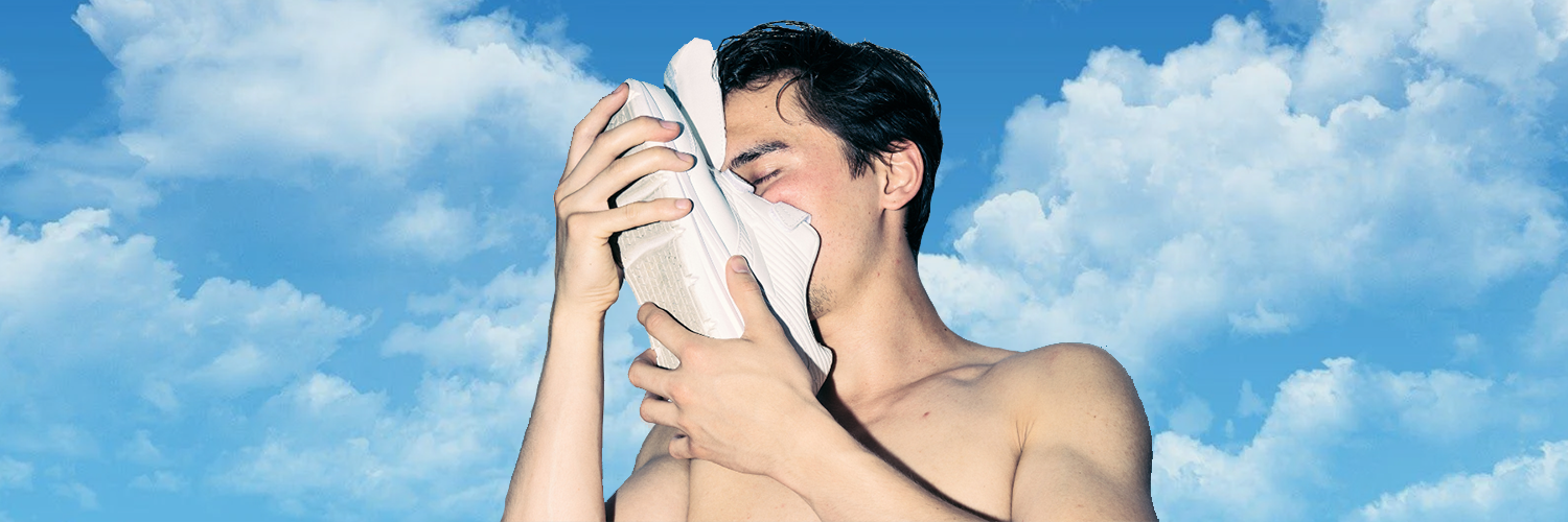 A shirtless boy sniffs a white shoe holding it to his face. The background is a clear blue sky with fluffy white clouds.