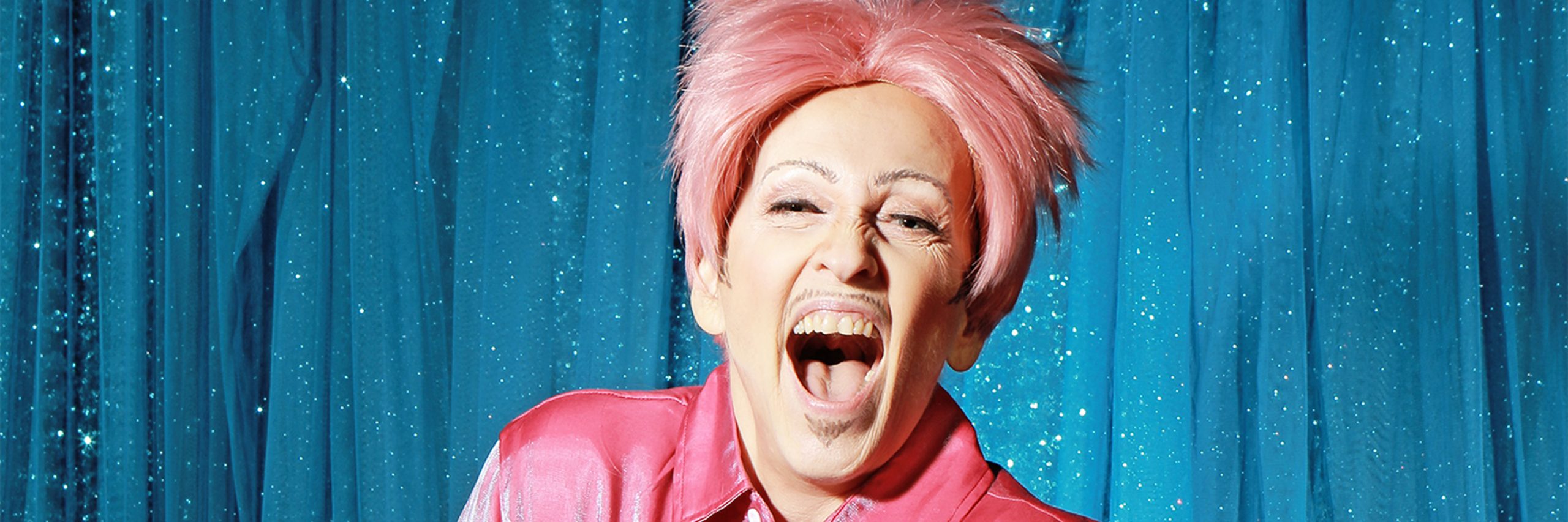 A drag king wearing a hot pink satin shirt and a pastel pink wig is laughing mouth open at the camera. With a sparkly blue curtain background.
